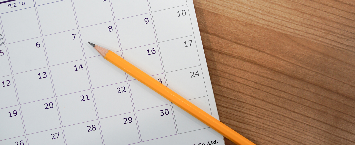 A close-up of a pencil resting against a calendar against a wood background.